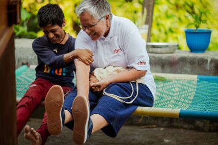 Eine Frau mit grauem Kurzhaarschnitt und ein Teenagerjunge sitzen lachend nebeneinander auf einer Liege mit bunter, gewebter Bespannung.