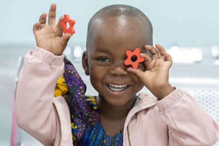 Ein kleines Mädchen lacht und hält eine rote Holz- oder Plastik-Blume vor sein linkes Auge. In der rechten Hand hält es eine zweite Blume hoch.