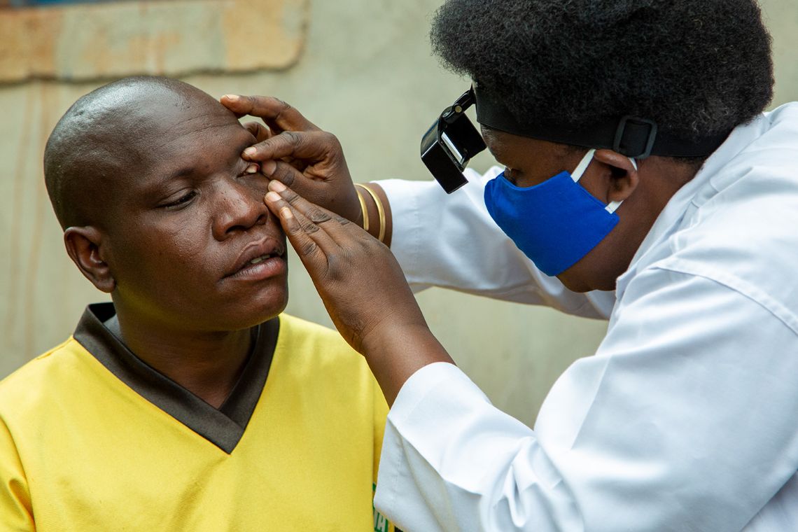 Eine Frau im weißen Kittel untersucht die Augen eines jungen Mannes im gelben T-Shirt.