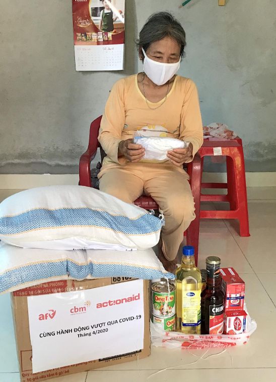 Eine Frau aus Asien sitzt vor einem Paket, auf dem zwei Säcke liegen. Daneben stehen Flaschen mit Öl und andere Lebensmittel.
